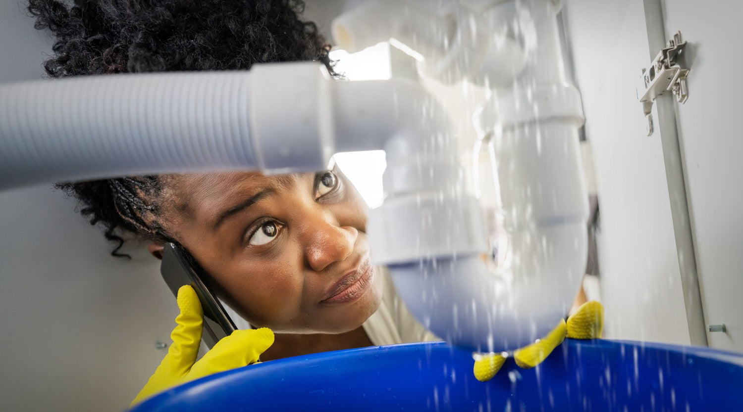 A woman looking under the sink at a hidden leak dripping into a bucket while on the phone with a plumber