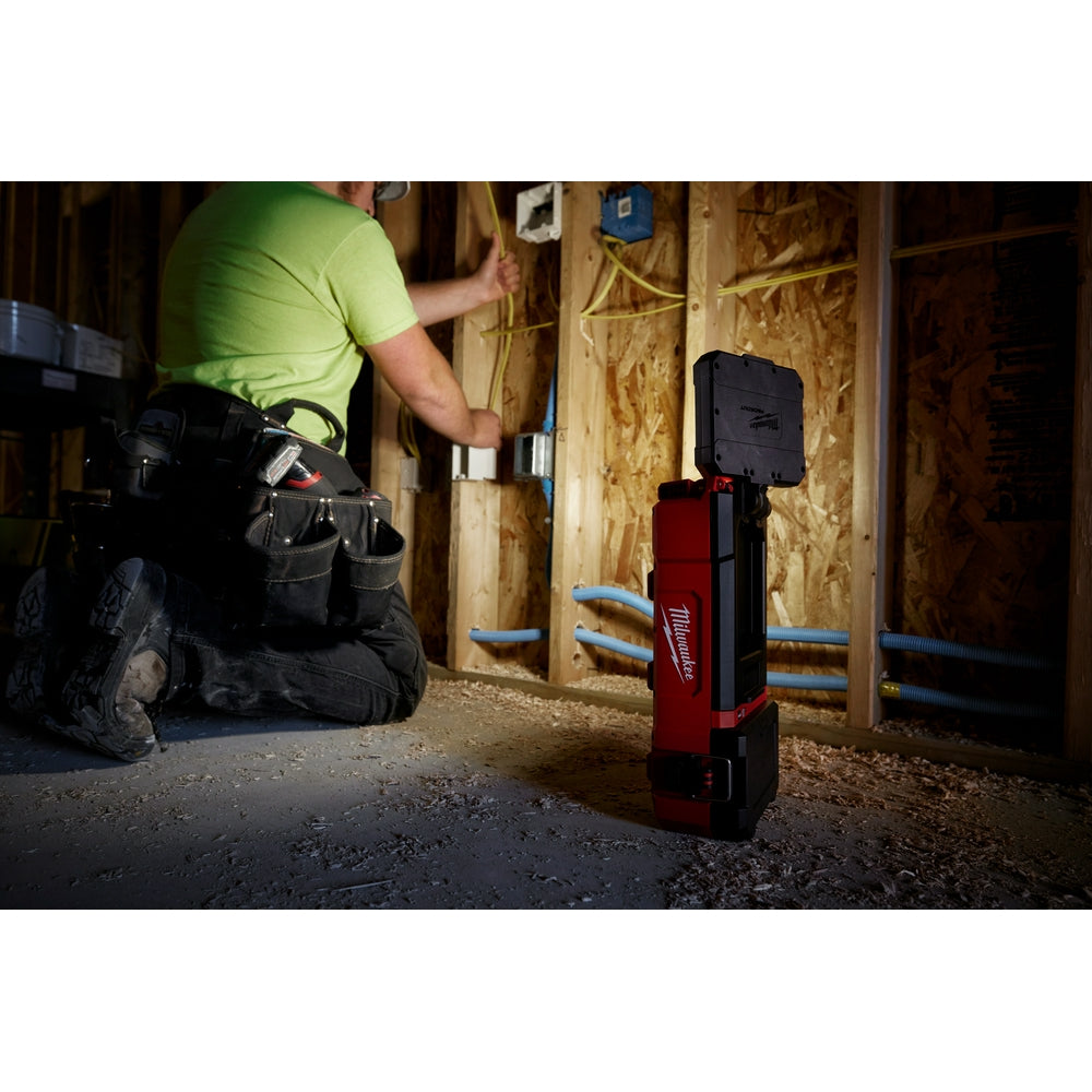 Red and black Milwaukee M12 PACKOUT flood light standing upright on a work site floor, illuminating a construction area.
