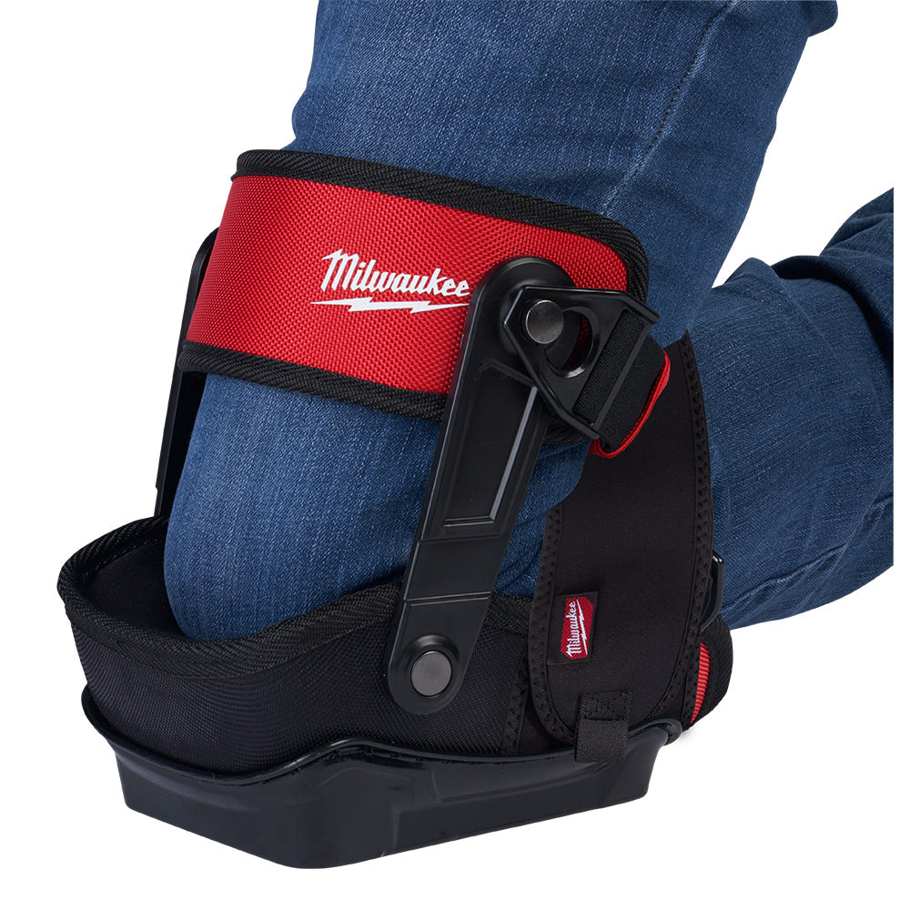 Red and black Milwaukee knee pad with stabilizer straps worn on kneeling leg over jeans.