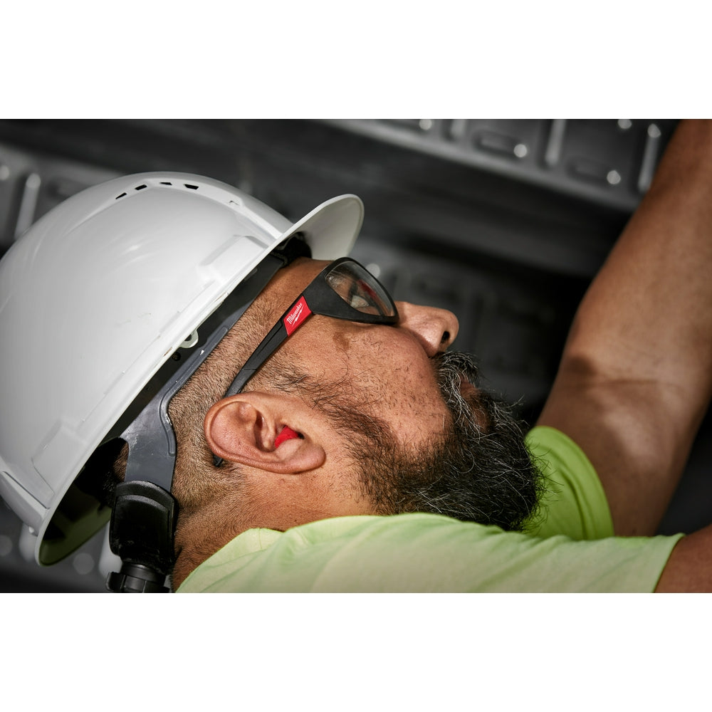 Red foam ear plugs inserted in a worker's ear wearing a white hard hat and safety glasses.