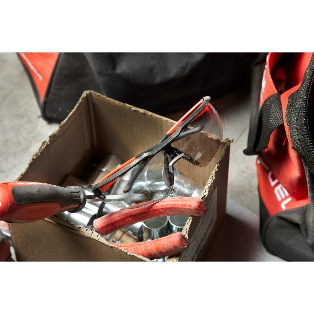 Safety glasses with gray lenses and red/black frames resting in a cardboard box next to work gloves.