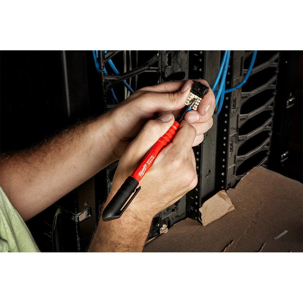 Red and black multi-pick tool with metal head held in hand near electrical equipment.
