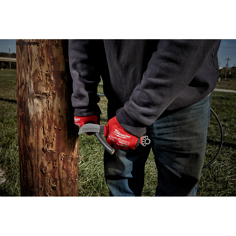 Red Milwaukee lineman knife with blunt hawkbill blade and multi-tool ring attachment, held against wooden utility pole.