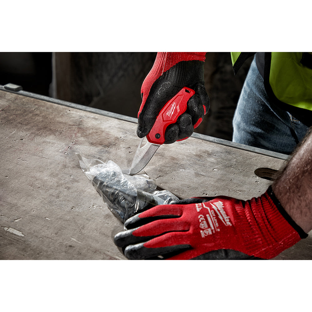 Red and black compact folding knife with blade extended, held in gloved hand cutting through plastic wrap on concrete surface