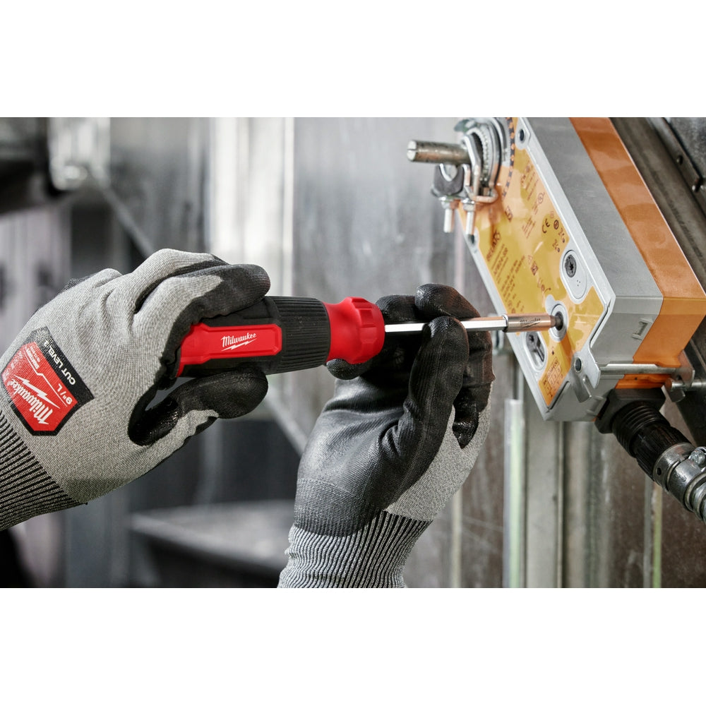 Red and black Milwaukee multi-bit screwdriver with hex handle held in gloved hands during use on metal surface.