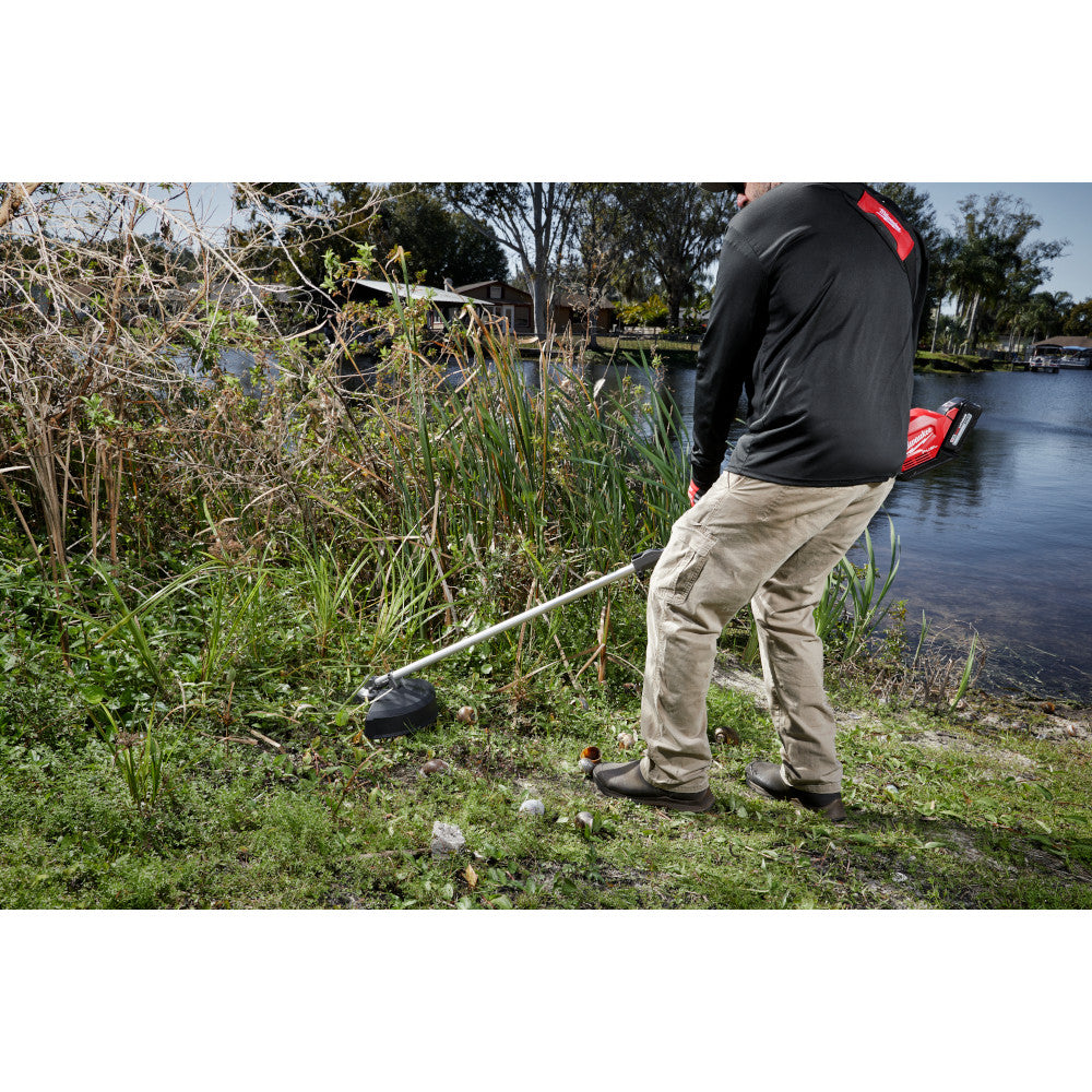 Person using Milwaukee brush cutter blade on overgrown vegetation near water.