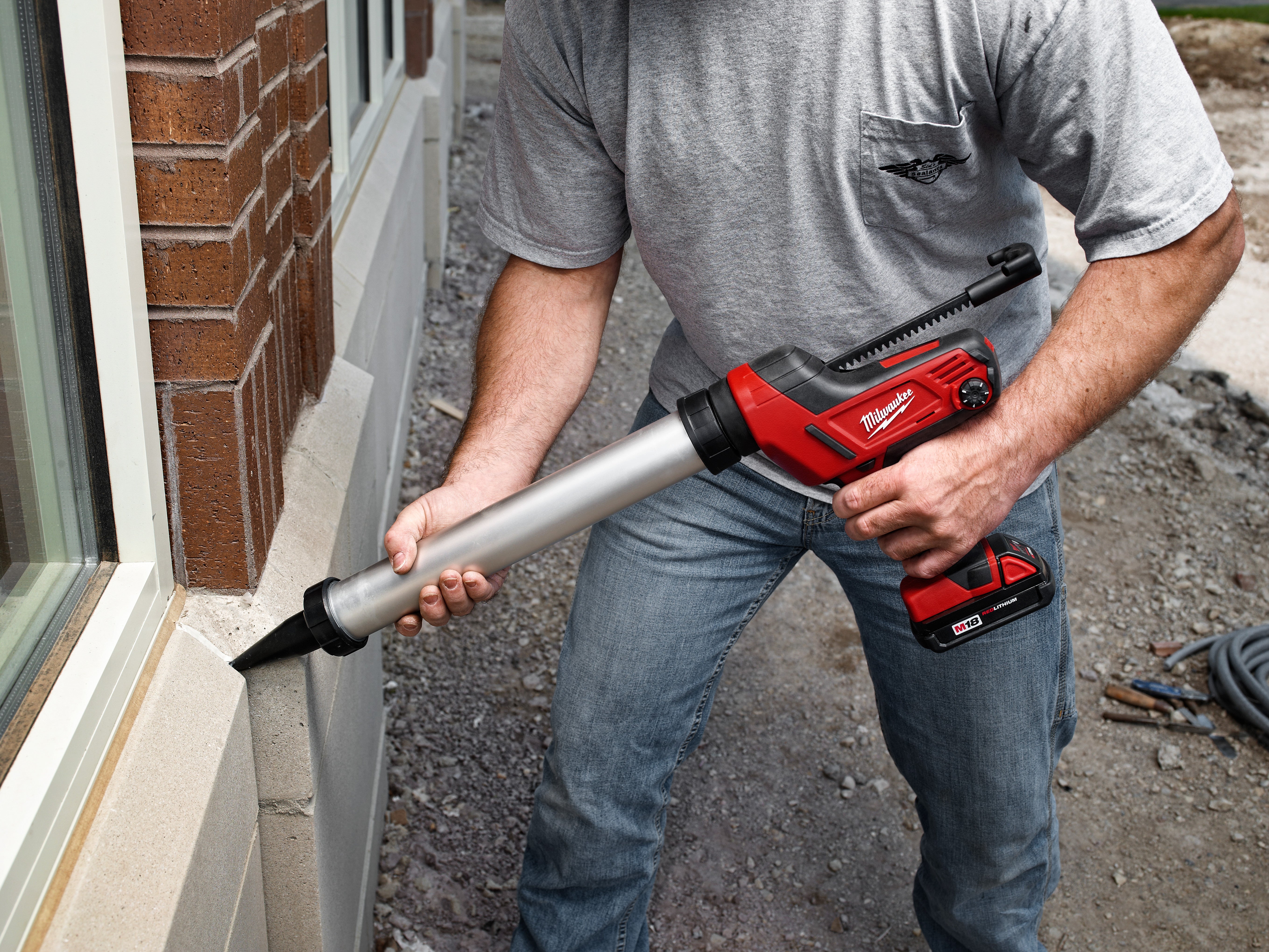 Person holding red and black caulking gun with aluminum barrel attachment against brick wall.