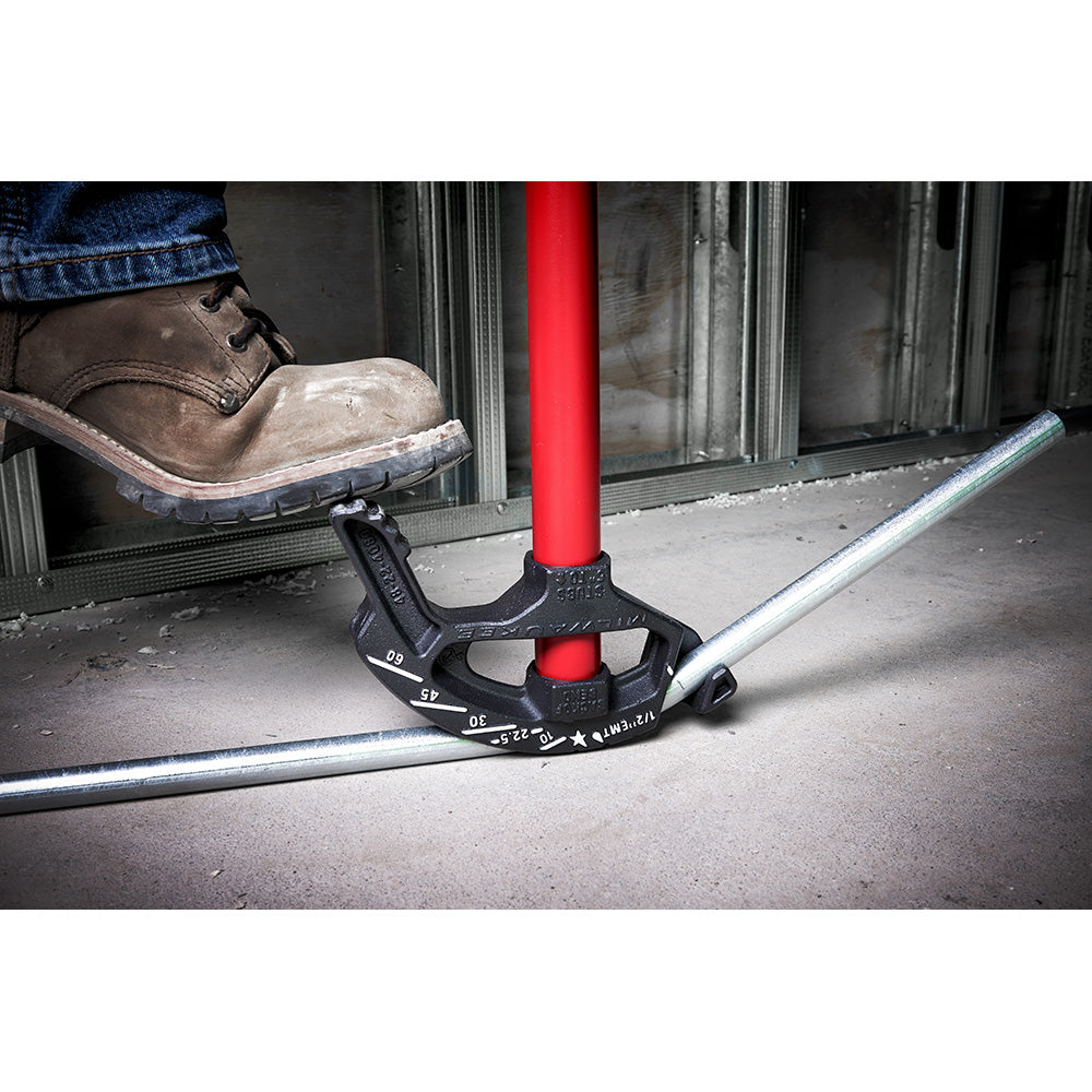 Black iron conduit bender with red handle gripping a metal pipe, positioned on concrete with a work boot visible.