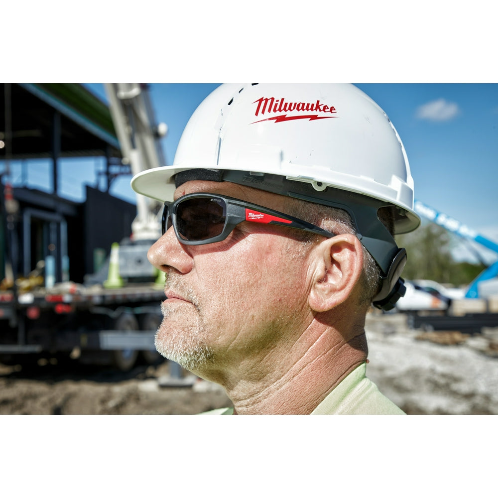 Tinted safety glasses with dark lenses and red accents worn on construction site by person in hard hat.