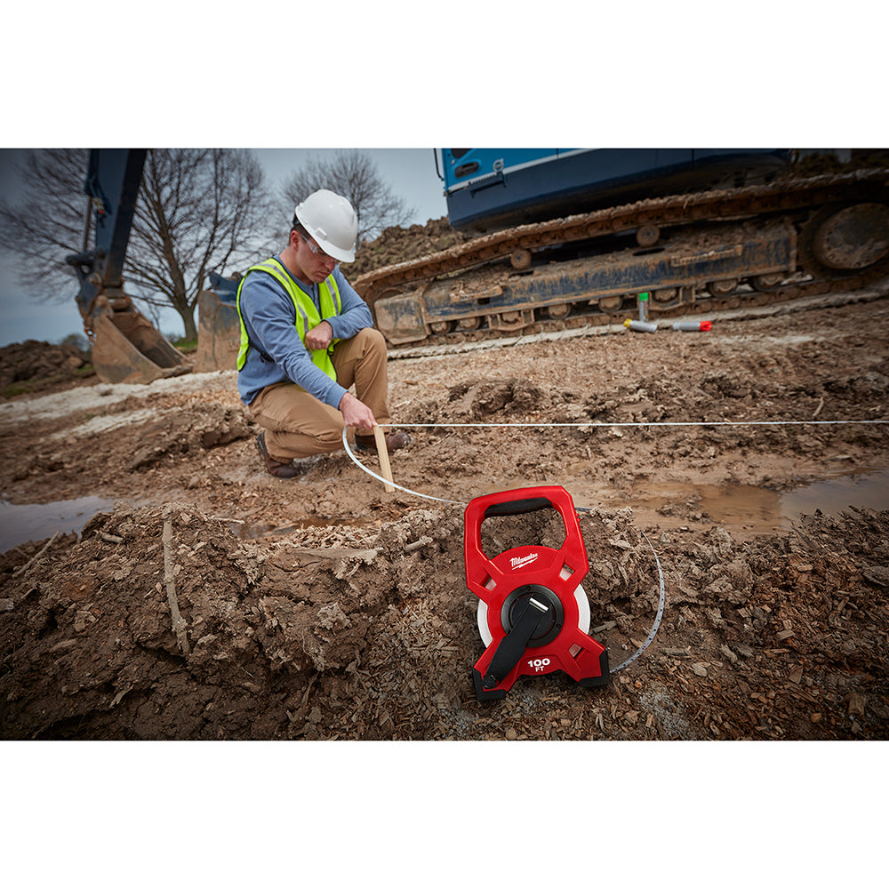 Red and white fiberglass open reel tape measure on dirt ground with worker holding extended tape.