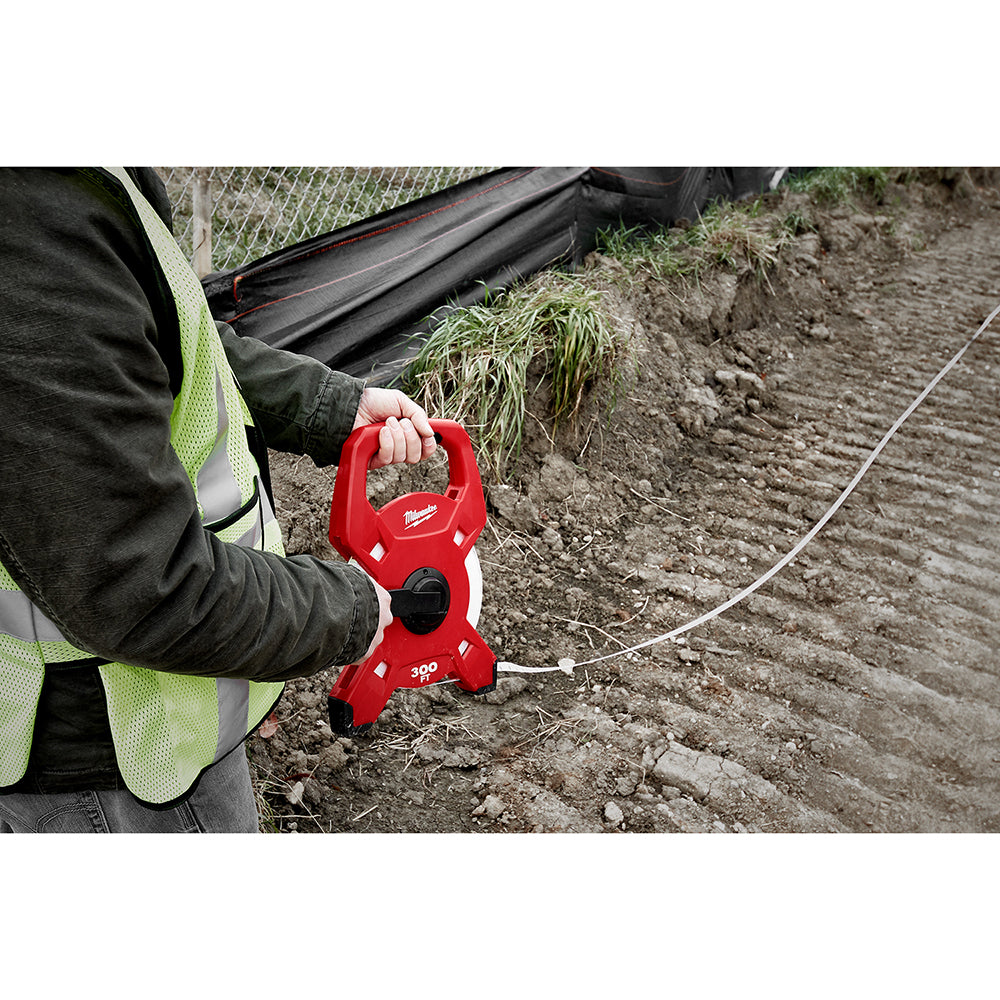 Red Milwaukee fiberglass tape reel with 300' marking held in worker's hand on job site.