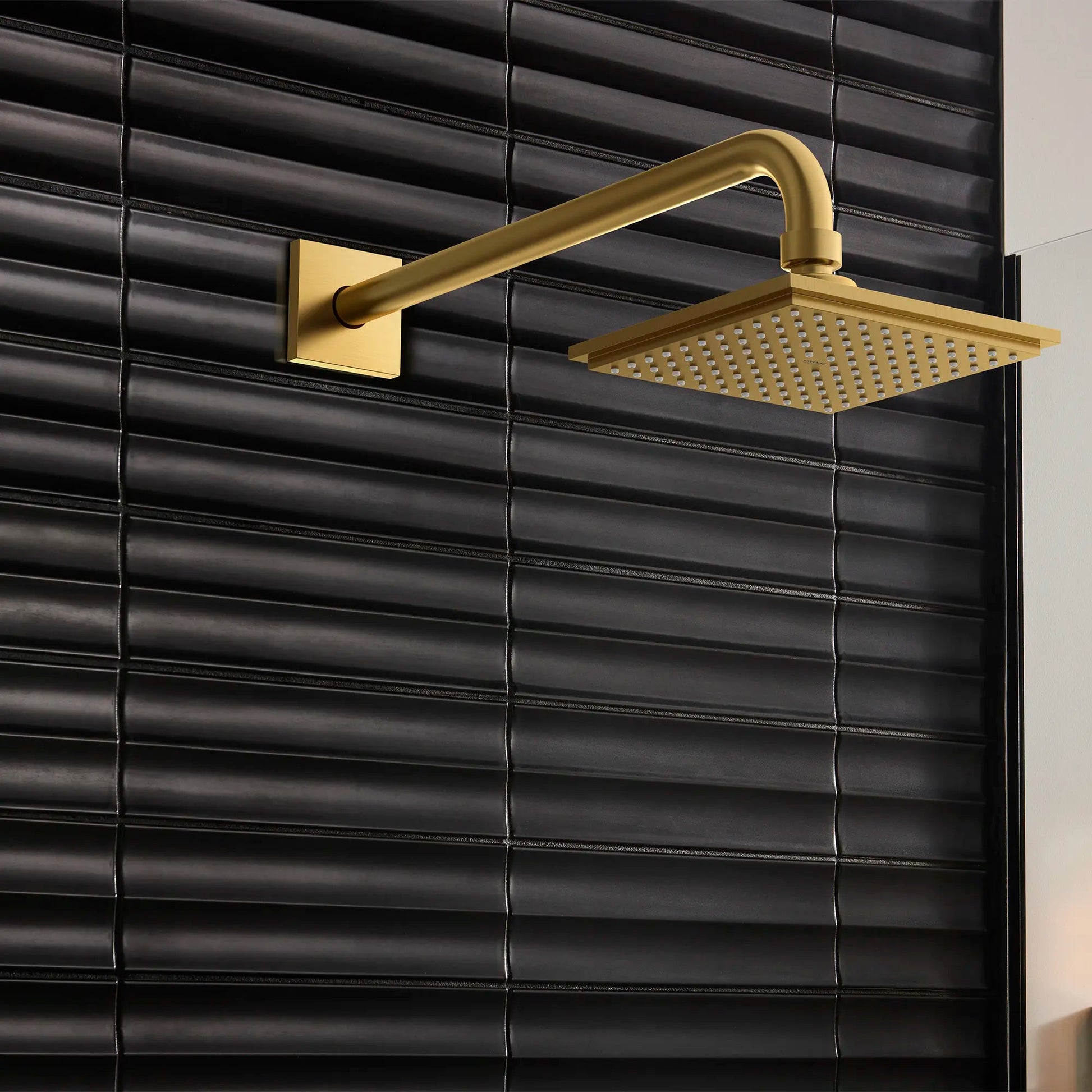 Gold square shower head with perforated face mounted on curved arm and square wall plate against black tile.