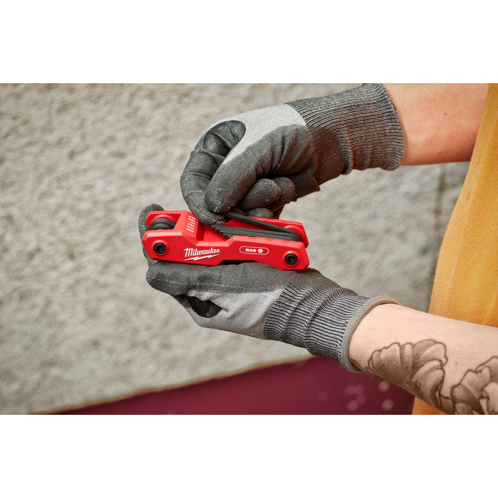 Red Milwaukee folding hex key set held in gloved hands, showing compact closed position with SAE marking visible.