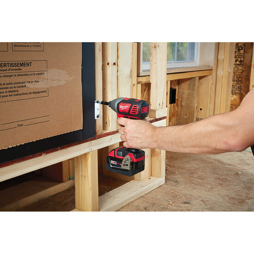 Milwaukee M18 impact driver with red and black body being held by a person's hand driving a screw into wood framing.