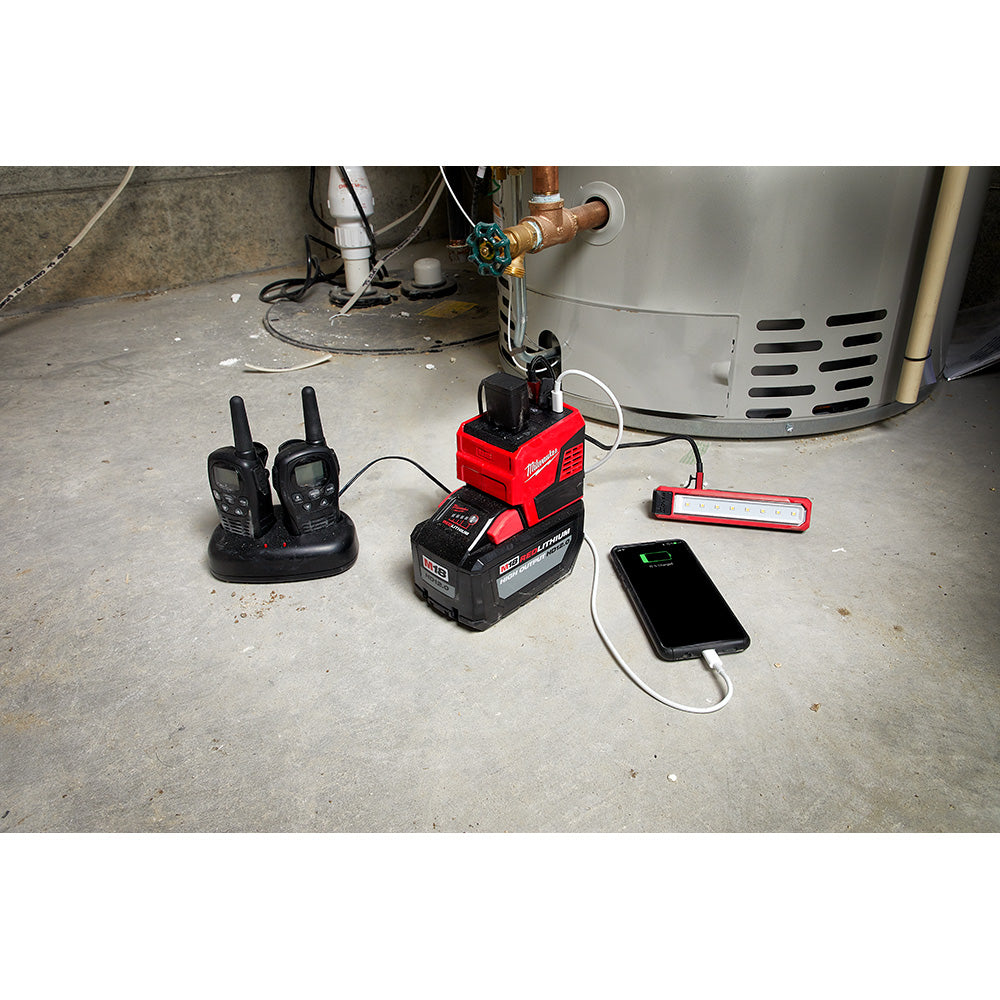 Milwaukee M18 power supply with red and black housing charging a smartphone and powering a work light and radios on concrete 