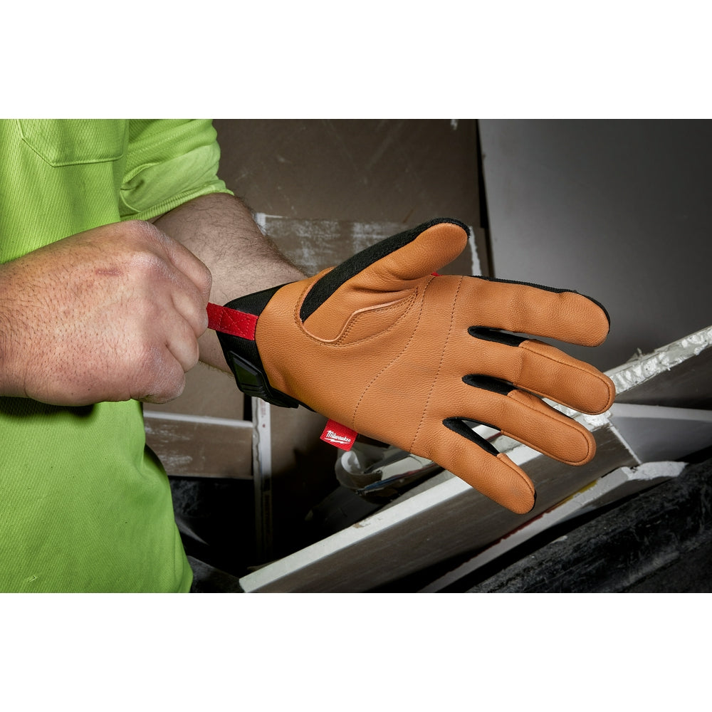Tan leather work glove with black fingers and red wrist strap on person's hand near metal frame.