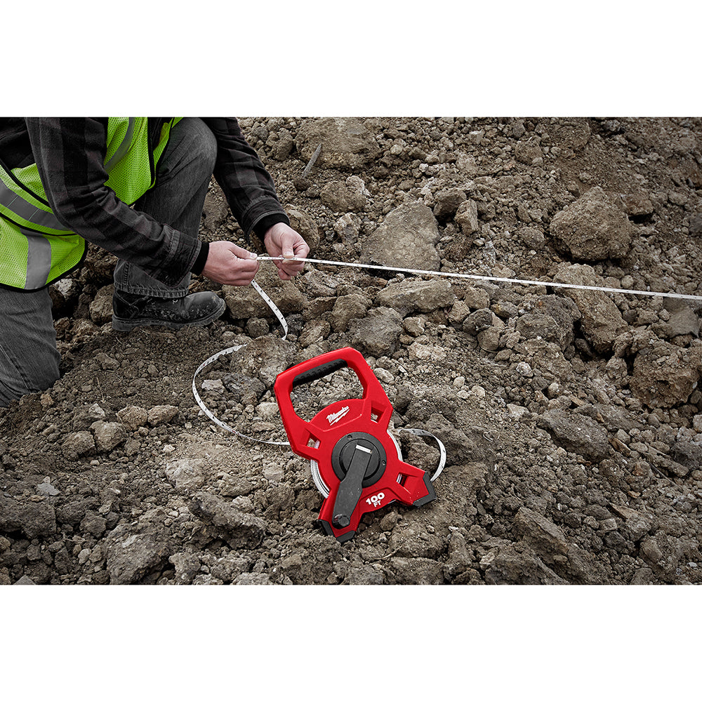 Red open reel tape measure with fiberglass tape extended across dirt ground, held by worker in safety vest.
