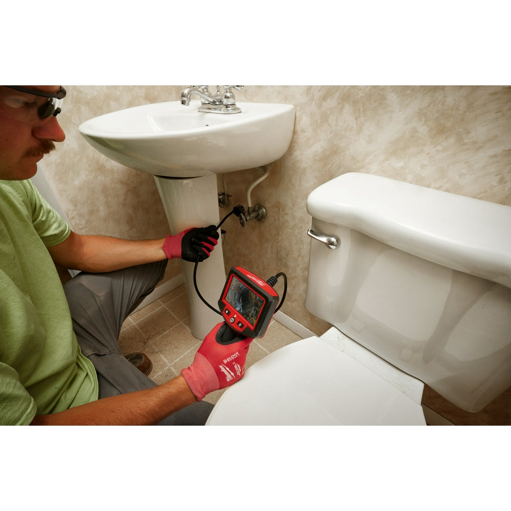 Red and black inspection camera with display screen being used under a bathroom sink by a person wearing gloves.