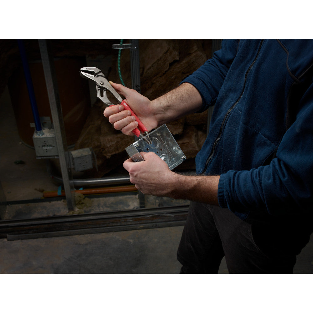 Straight-jaw pliers with red handles held in use gripping metal material in a workshop.