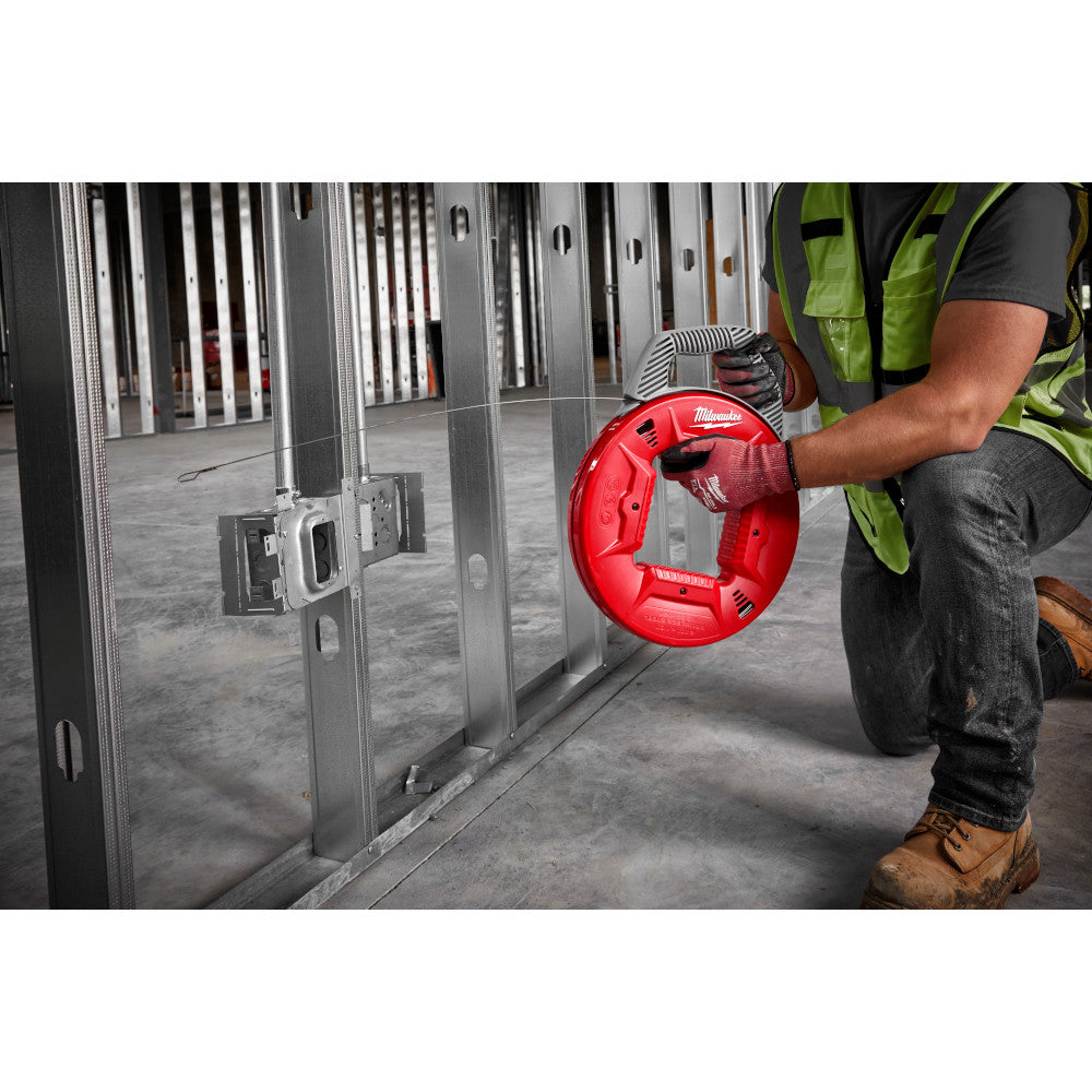 Worker holding red fish tape reel with stainless steel tape extended toward metal framing in construction setting.