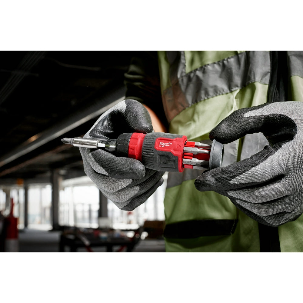 Red and black compact multi-bit screwdriver with ratcheting head held in gloved hands in a workshop setting.