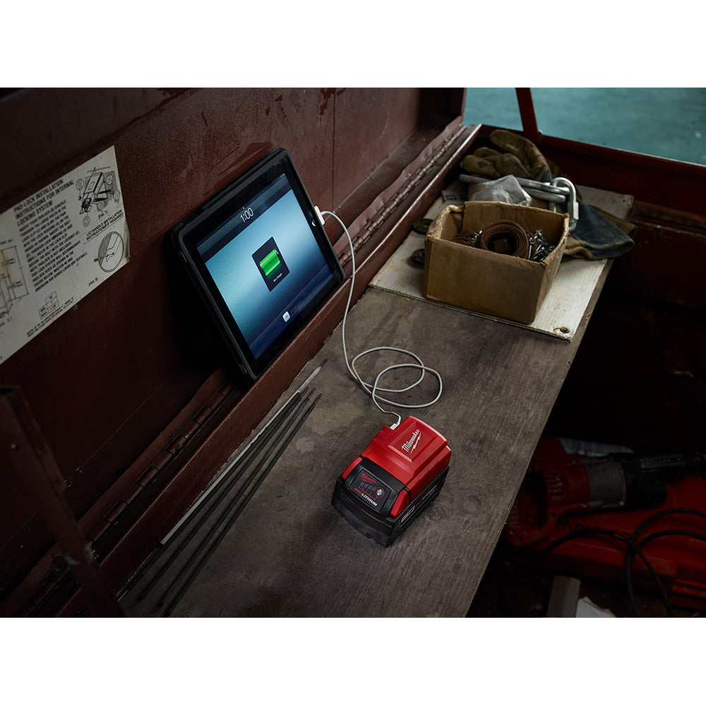 Red and black Milwaukee M18 Power Source connected by cable to tablet displaying charging status on work surface.