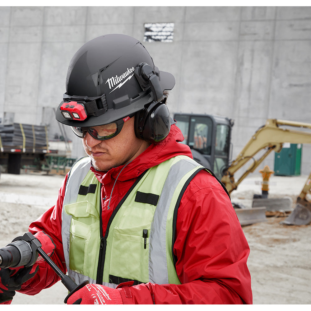 Green full brim hard hat with ratcheting suspension, front-mounted red light, and ear muff attachments worn on construction s