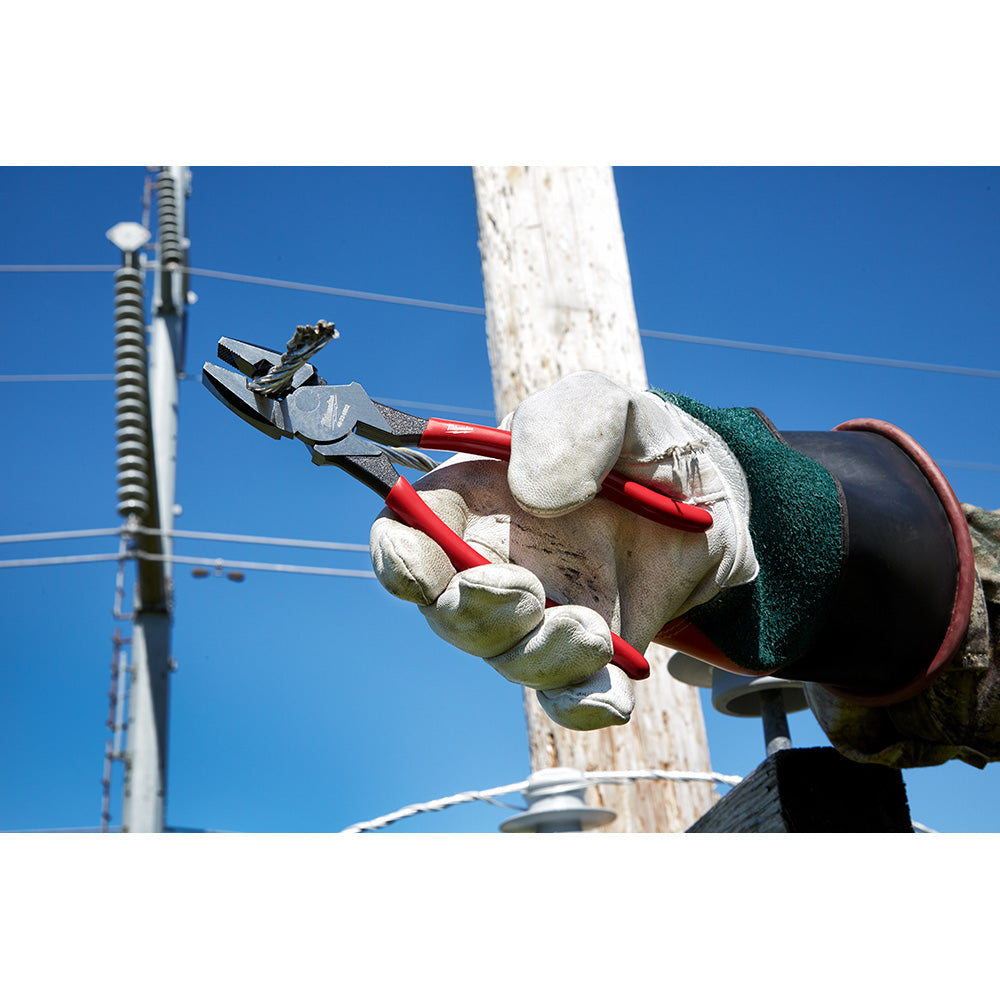 Linesman's pliers with red handles and dipped grip held in a gloved hand near electrical utility lines and pole.