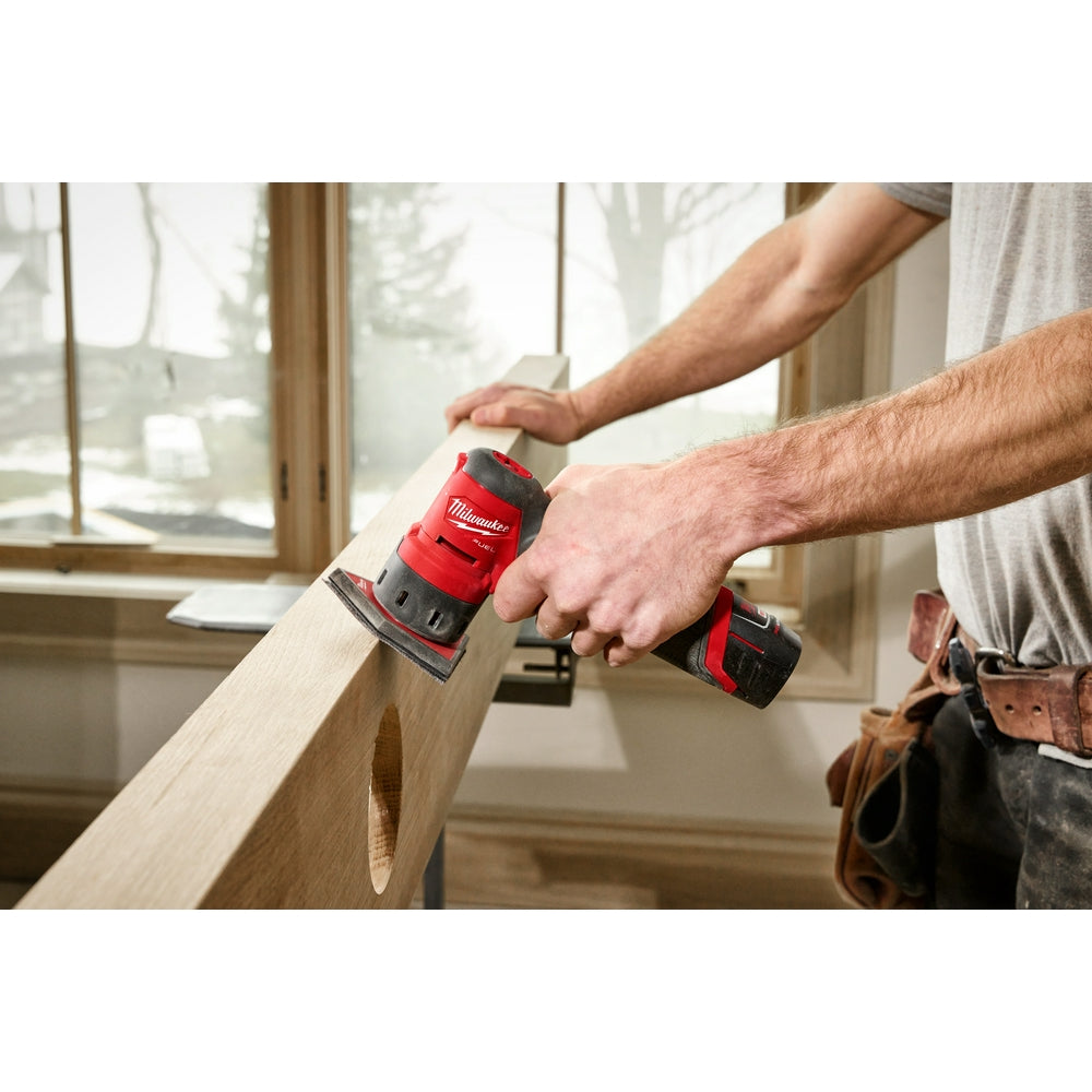Red and black Milwaukee M12 orbital detail sander with rectangular sanding pad being used on wood trim.