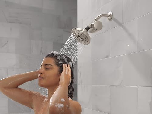 Brushed nickel shower head and arm mounted on white tile wall with water flowing down onto person's head.
