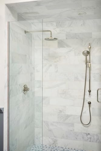 Round brass showerhead mounted on ceiling with hand shower and controls on marble tile bathroom wall.