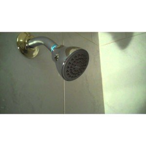 Round black shower head with brass wall mount and adjustment valve on tiled wall.