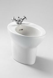 White ceramic bidet with oval rim and chrome cross-handle faucet mounted on top rim, center drain opening visible.