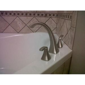 Bronze two-handle deck mount Roman tub faucet with curved spout and cone-shaped handles installed on white bathtub.