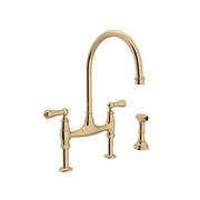 Unlacquered brass bridge kitchen faucet with high arched spout, dual handles, and separate side spray attachment.