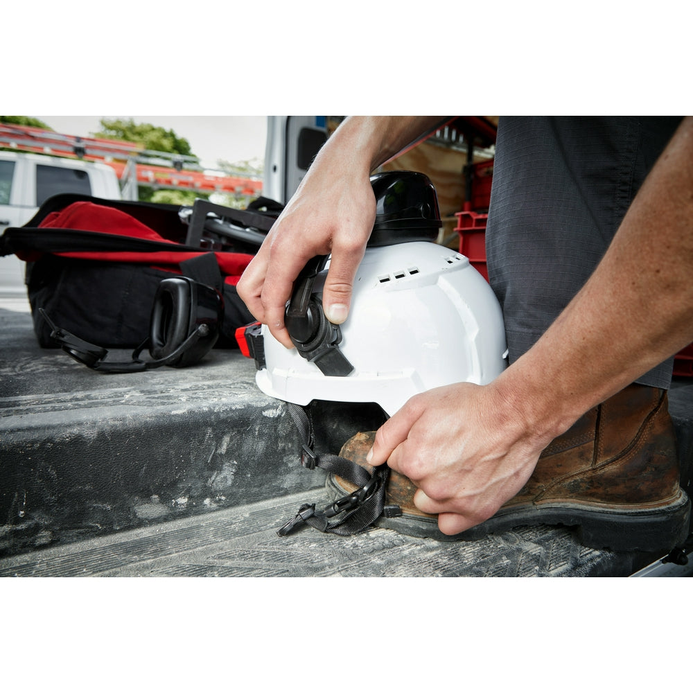 Gray vented safety helmet with black chin strap and ventilation slots, held in hands on work site.