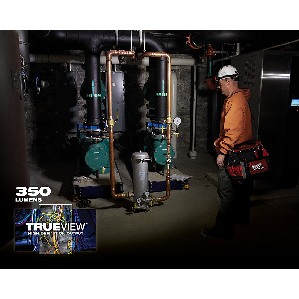 Worker wearing white hard hat and orange jacket with Milwaukee headlamp illuminating industrial basement equipment.
