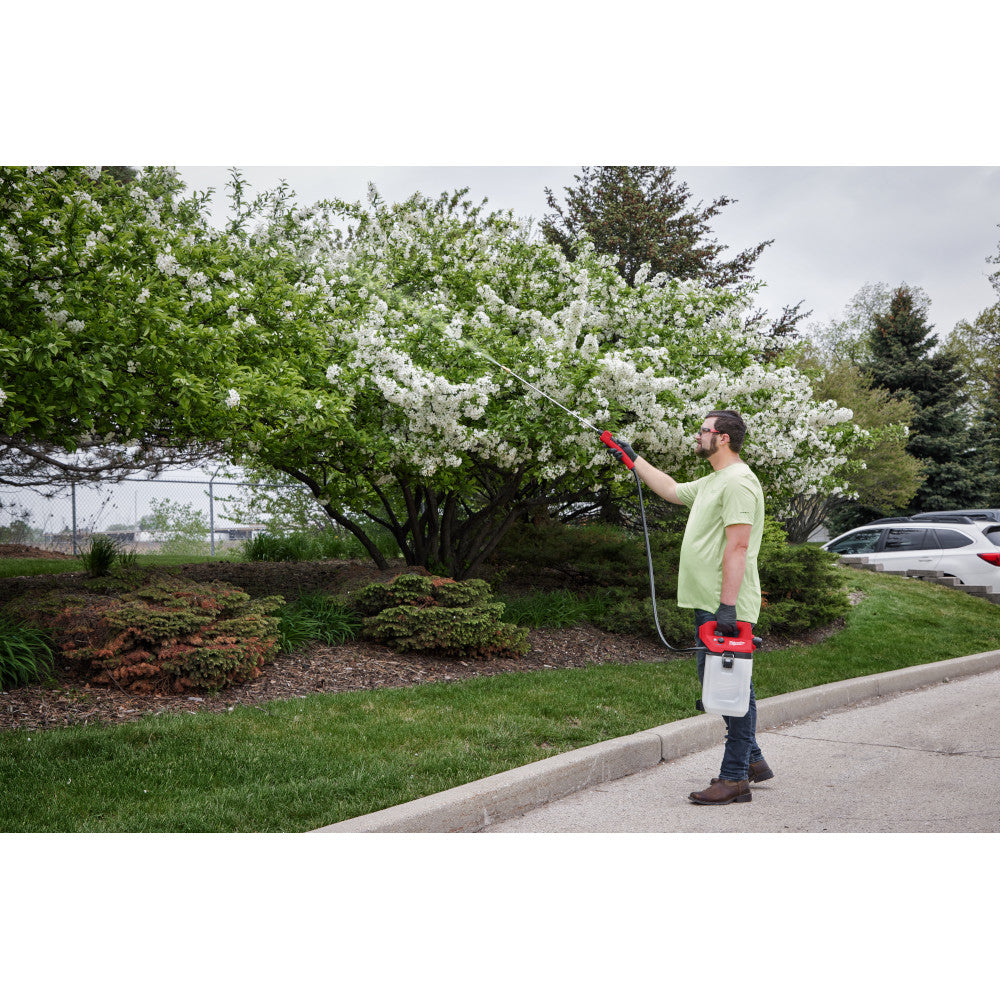Red and white handheld sprayer tank with shoulder strap being used to spray flowering shrubs outdoors.