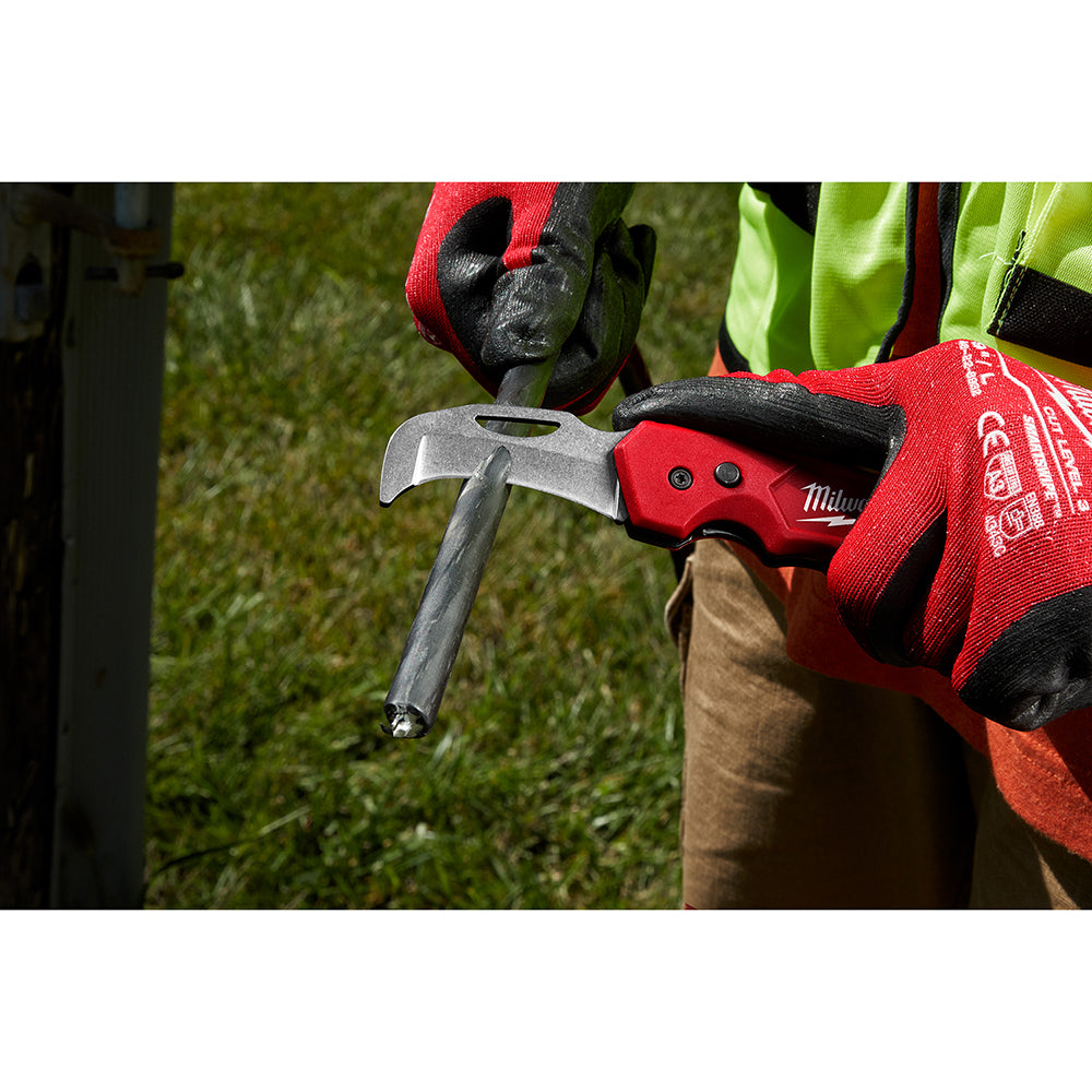 Red and black folding knife with curved blade held in gloved hand outdoors.