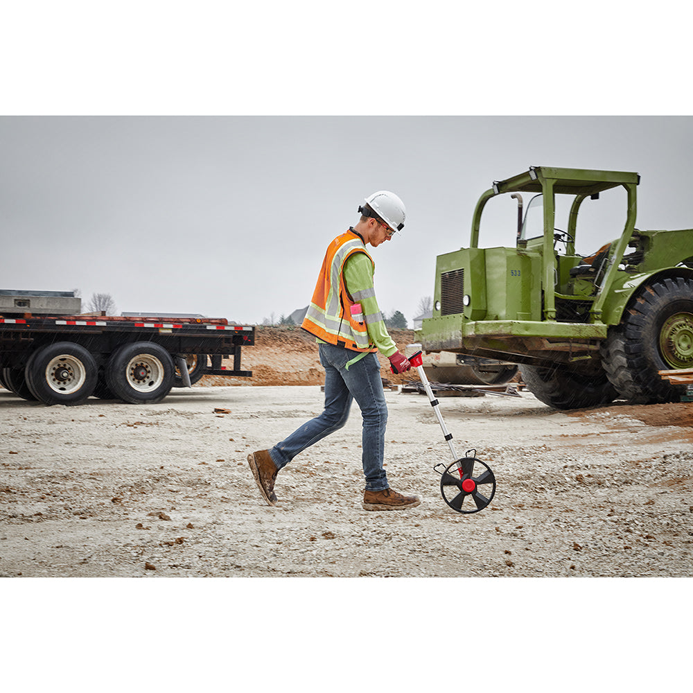 Worker in safety gear rolling a digital measuring wheel with red and black wheel on gravel surface.