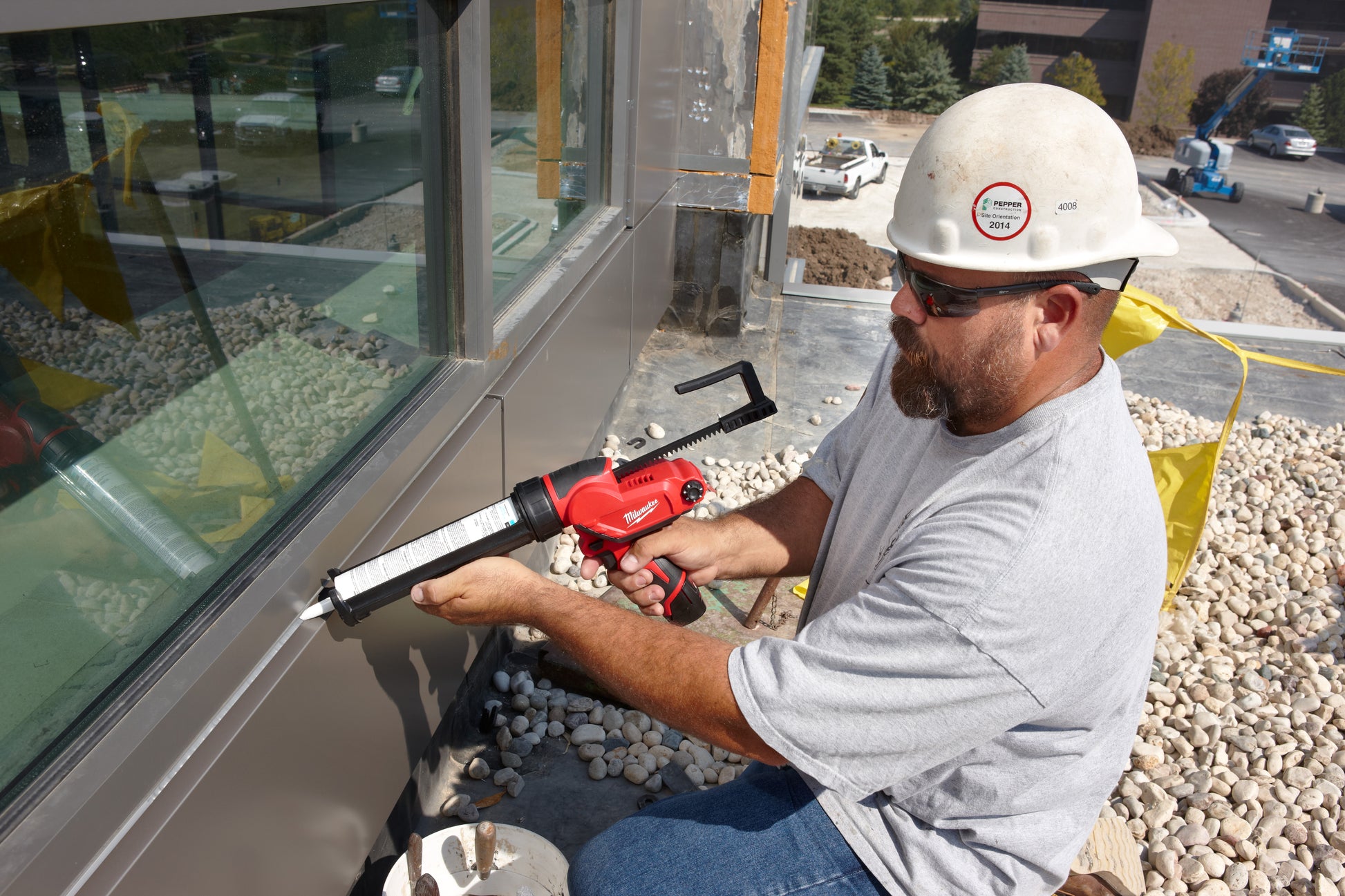 Worker holding red and black caulking gun with aluminum barrel attachment outdoors.