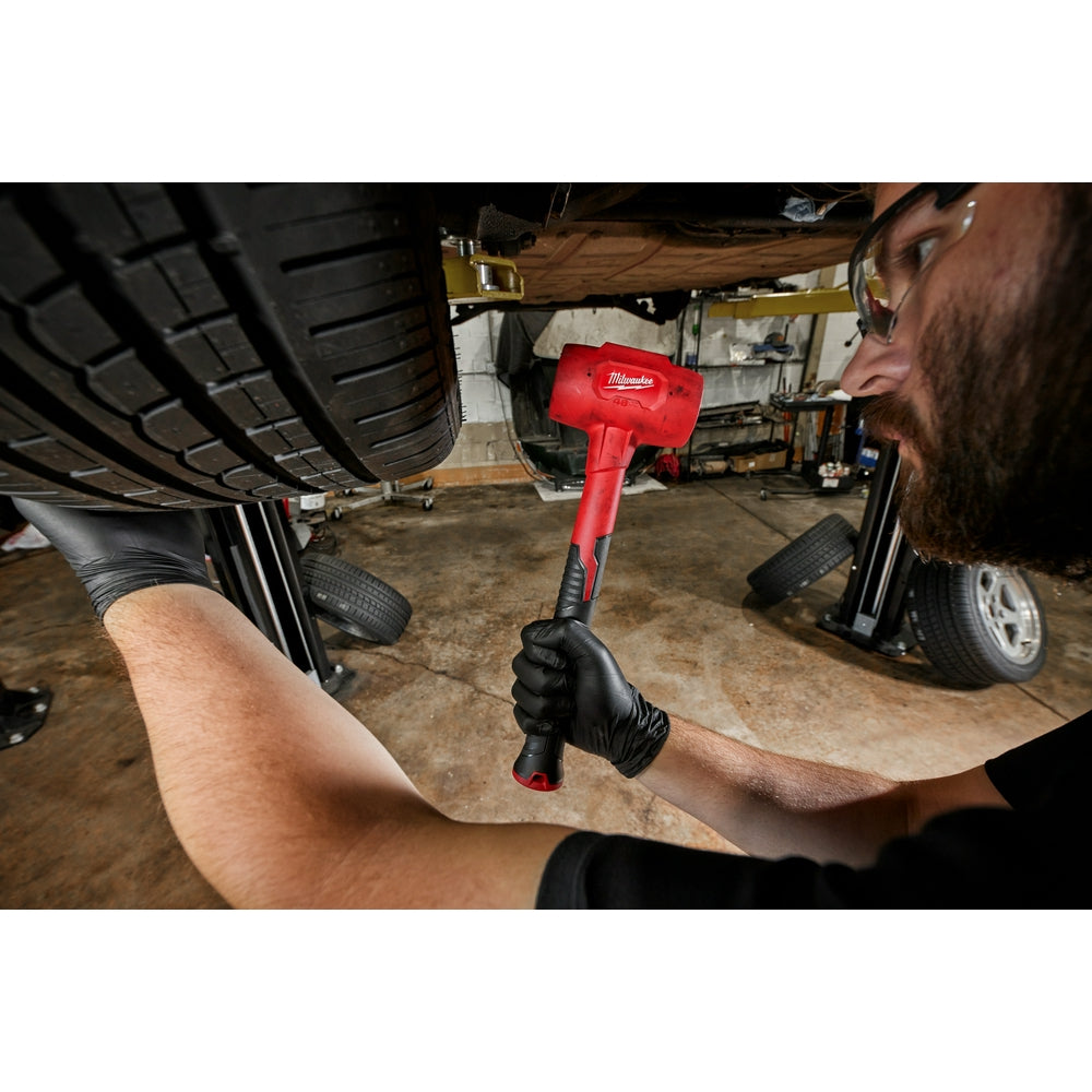 Red and black Milwaukee dead blow hammer with cylindrical head held by person wearing gloves in workshop setting.