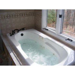 Brushed nickel two-handle deck mount roman tub faucet with handheld showerhead installed on white bathtub.
