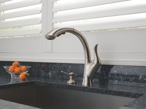 Stainless steel soap and lotion dispenser with curved spout mounted on dark countertop next to kitchen sink.
