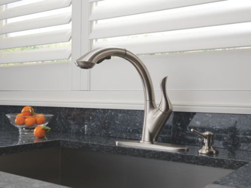 Stainless steel soap dispenser with curved spout mounted next to kitchen faucet on dark granite countertop.