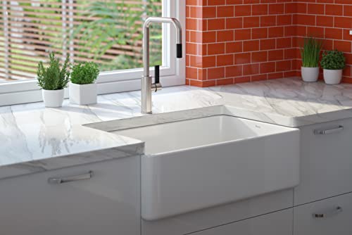White fireclay single bowl farmhouse sink with apron front installed in gray cabinetry beneath a marble countertop.