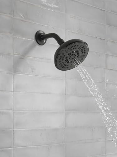 Matte black circular shower head with curved arm mounted on white subway tile wall, water streaming downward.