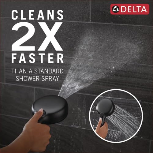 Matte black hand shower held in hand spraying water against tiled wall surface.
