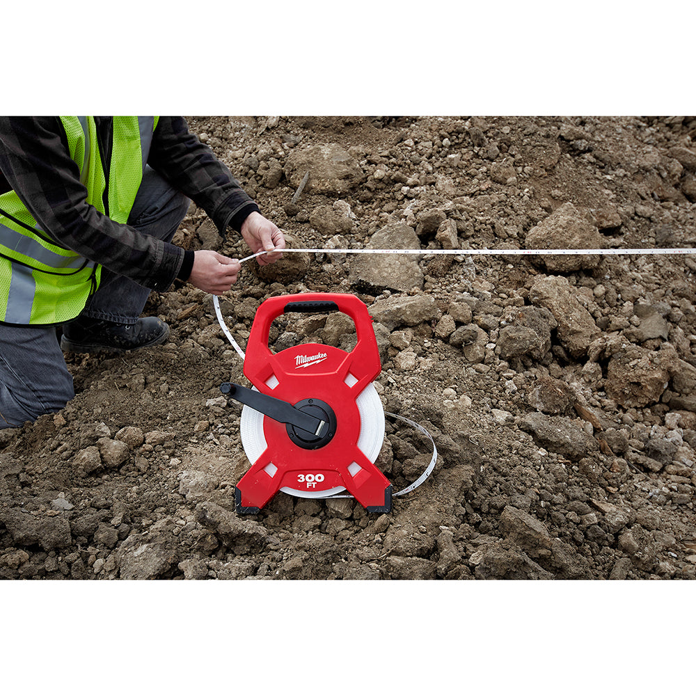 Red and white Milwaukee open reel fiberglass tape measure with worker in safety vest measuring distance on dirt ground.