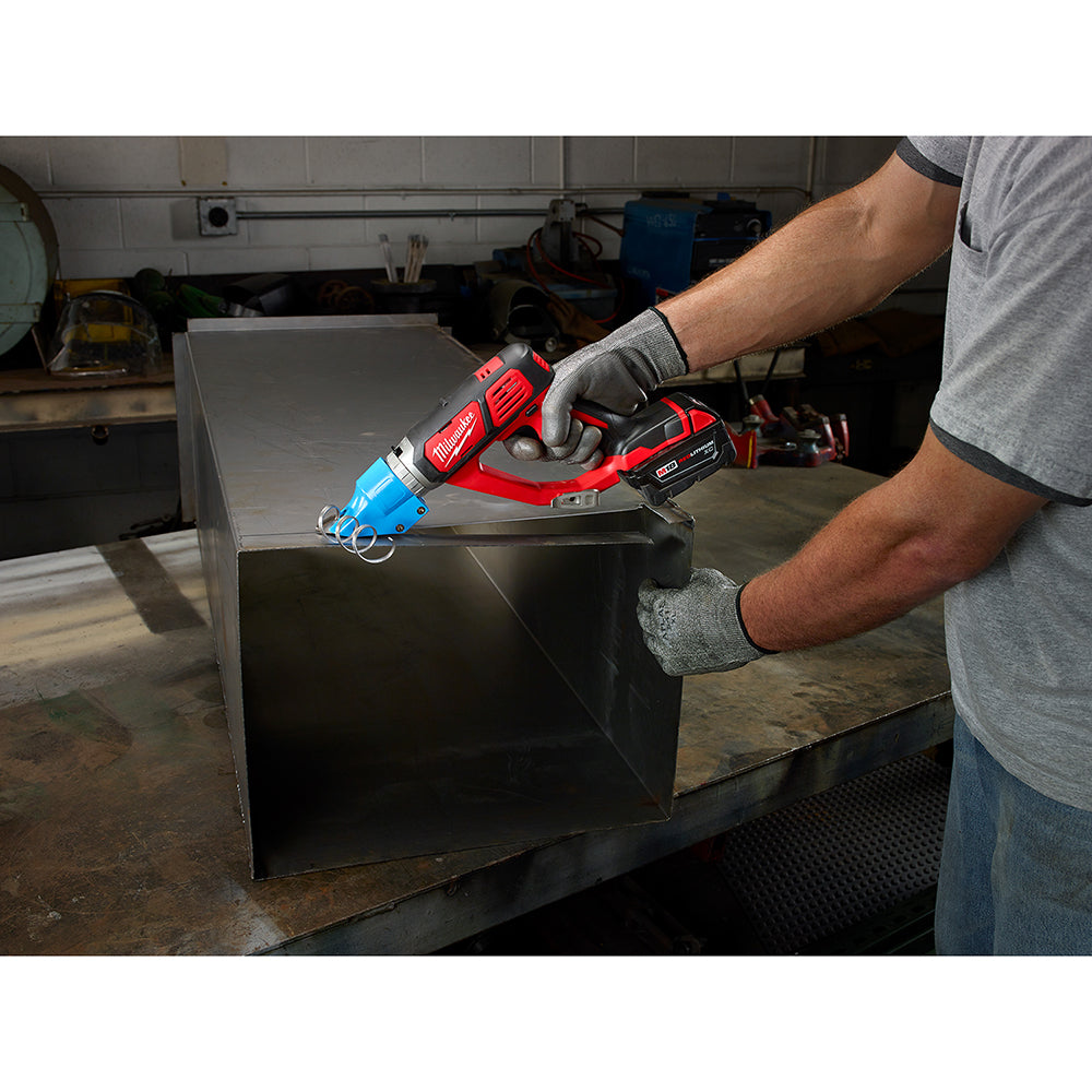Red and blue Milwaukee cordless shear with battery being held above a metal sheet in a workshop.
